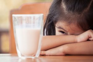 Menina debruçada na mesa com um cara sofrida olhando para um copo de leite por ter alergia à proteína do leite de vaca