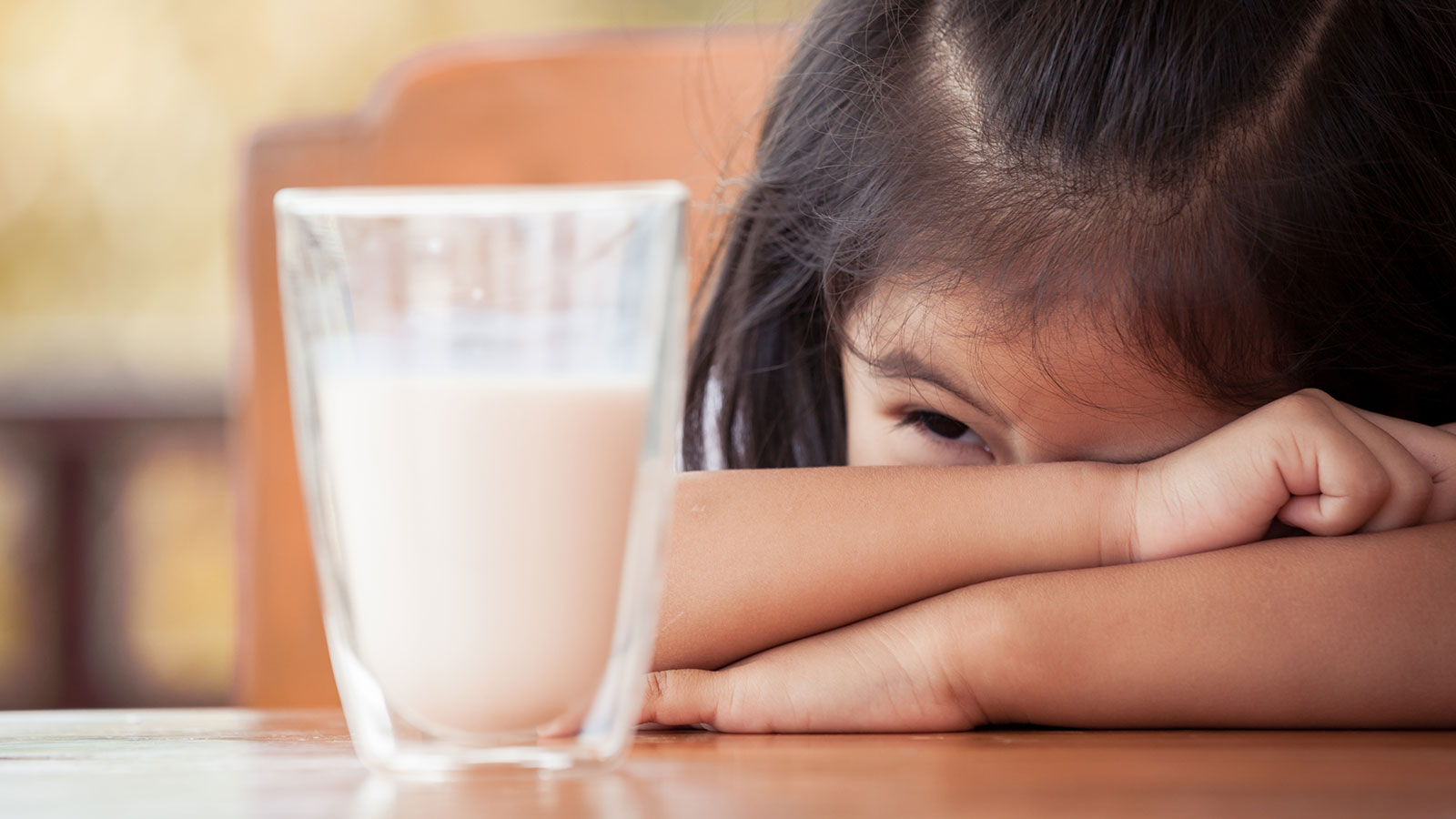 Menina debruçada na mesa com um cara sofrida olhando para um copo de leite por ter alergia à proteína do leite de vaca