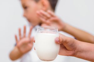 Close de uma mão com um copo de leite enquanto, ao fundo, um garoto desfocado rejeita o alimento por ter intolerância à lactose