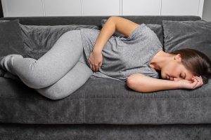 Adolescente deitada de olhos fechados em um sofá com um das mãos na barriga e outra no rosto indicando que está sofrendo de alguma dor proveniente de doença inflamatória intestinal