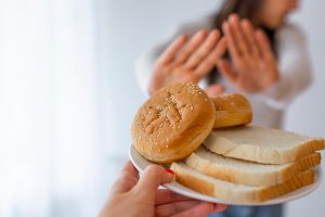 Mão segurando um prato com pães no primeiro plano e, no segundo plano, uma mulher desfocada recusa a comida por ter doença celíaca